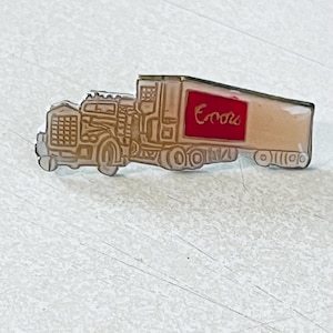 May include: A pin shaped like a semi-truck, featuring a beige trailer with a red rectangle and the word "Encore" in red script. The truck's cab is detailed in a lighter beige color with gold trim.