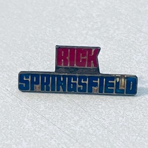 Puede incluir: Un pin rectangular con el nombre "RICK" en letras rojas sobre el nombre "SPRINGFIELD" en letras azules. El pin tiene un borde plateado y está sobre un fondo gris claro.