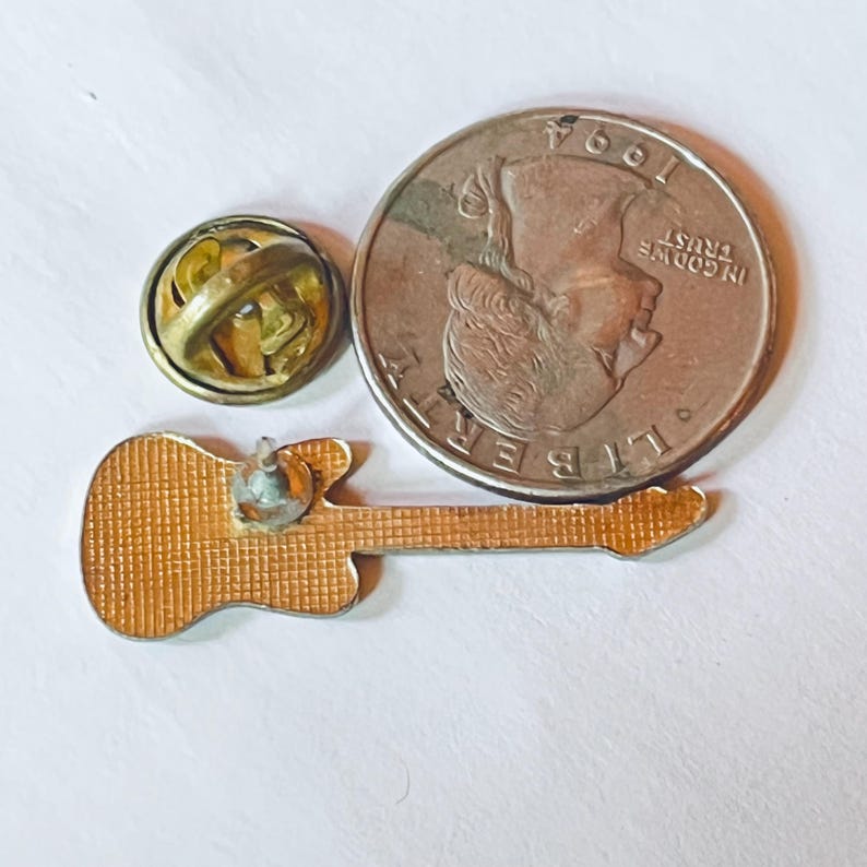 Op de afbeelding: Een goudkleurige metalen reversspeld in de vorm van een gitaar, getoond vanaf de achterkant, met een gestructureerd rasterpatroon en een enkele pin. Een ronde metalen sluiting ligt ernaast, samen met een Amerikaanse kwartje uit 1994 voor schaal.