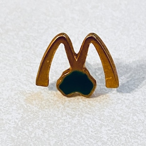May include: A gold-colored pin featuring the iconic "M" logo with a dark green enamel fill at the base. The pin has a classic design, with the two arches connected at the bottom. The pin is likely a collectible or promotional item.