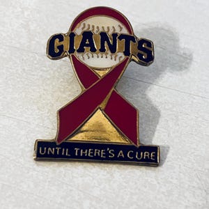 May include: A baseball-themed pin featuring the word "GIANTS" in navy blue lettering above a baseball. A burgundy ribbon design is below the baseball. The pin includes the text "UNTIL THERE'S A CURE" in navy blue.