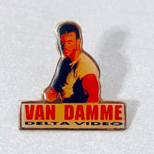 May include: A gold-coloured pin featuring a photo of Jean-Claude Van Damme in a fighting pose. The pin has the text "Van Damme" and "Delta Video" in red letters.
