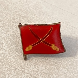 Puede incluir: Un pequeño pin rectangular con un diseño de bandera roja con dos espadas cruzadas. Las espadas tienen empuñaduras y hojas doradas. El pin tiene un borde plateado y un pequeño poste plateado para la fijación. La bandera está ligeramente curvada.