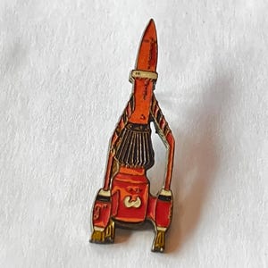 Puede incluir: Pin de cohete de estilo vintage, con una combinación de colores rojo y naranja. El pin presenta una parte superior puntiaguda, una sección media detallada con un diseño similar a una falda marrón y el número "3" en la parte inferior. Se aprecian detalles dorados.