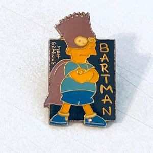 May include: Enamel pin featuring Bart Simpson as Bartman. The pin has a black background with the word "BARTMAN" in gold lettering. Bart is wearing a purple cape and mask, a blue shirt and shorts, and yellow shoes. The pin is signed "Matt Groening".