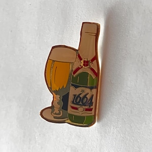 Puede incluir: Un pin dorado con un vaso de cerveza y una botella verde con el número "1664". El vaso está lleno de un líquido dorado, y la botella tiene detalles rojos y verdes.