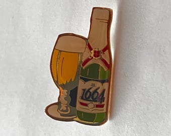 May include: A gold-coloured pin badge depicting a beer glass and a green bottle with the number "1664". The glass is filled with a golden liquid, and the bottle has red and green detailing.
