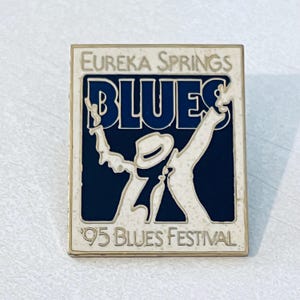 Op de afbeelding: Een rechthoekige pin met een gouden rand met de tekst "Eureka Springs Blues" boven een gestileerde afbeelding van een muzikant. Het woord "BLUES" staat in grote blauwe letters. De tekst "'95 Blues Festival" staat onderaan.