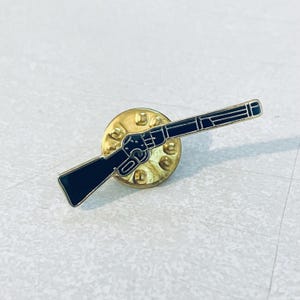 May include: A black rifle pin with a gold-coloured backing. The pin features a detailed outline of a rifle, with a black enamel finish. The pin is a small, decorative accessory, likely for a jacket or bag.