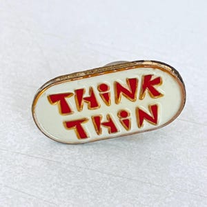 May include: An oval pin badge with the words "THINK THIN" in red lettering. The letters are outlined in gold against a white background. The badge has a gold-coloured border.