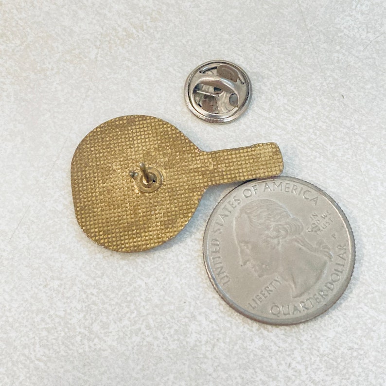 May include: Gold-toned metal pinback button featuring a stylized image of a table tennis bat. The back of the pin has a standard pinback closure. The pin is next to a US quarter for size reference.