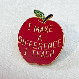 May include: A red enamel apple-shaped pin with gold trim and the text "I MAKE A DIFFERENCE I TEACH" in gold lettering. The apple has a green leaf and a black stem. A teacher appreciation gift.