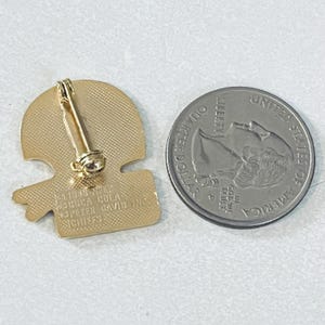 May include: A gold-colored pin with a football helmet design, featuring a pin back. Next to the pin is a United States quarter coin. The pin has text that reads "1990 NFL USA COLA PETER DAVID INC CHIEFS."