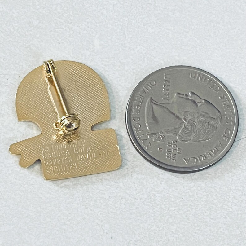 May include: A gold-colored pin with a football helmet design, featuring a pin back. Next to the pin is a United States quarter coin. The pin has text that reads "1990 NFL USA COLA PETER DAVID INC CHIEFS."