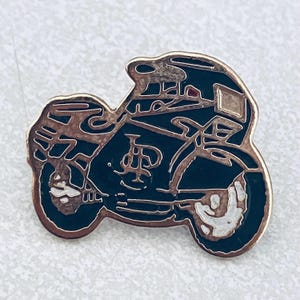 May include: A motorcycle pin with a black body and gold-colored trim. The pin features a detailed design of a motorcycle, including the wheels, seat, and handlebars. The pin is likely made of metal and has a shiny finish.