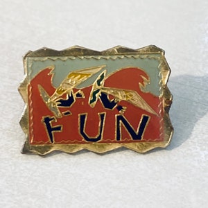 May include: A gold-colored pin with a red, white, and blue design. The pin features two stylized birds flying with the word "FUN" in black letters on a red background.