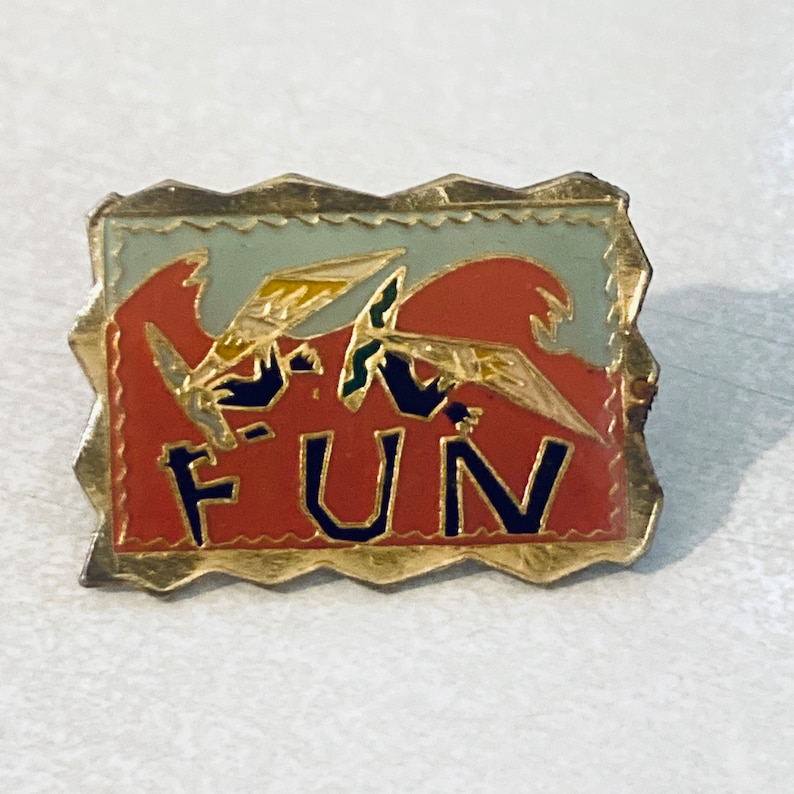 May include: A gold-colored pin with a red, white, and blue design. The pin features two stylized birds flying with the word "FUN" in black letters on a red background.