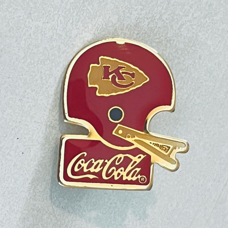 May include: A red and gold pin featuring a football helmet design with the Kansas City Chiefs logo and the Coca-Cola logo. The pin has a gold border and a black circle detail.