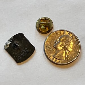 May include: A vintage metal pin with a rectangular shape, a gold-colored pin back, and a US quarter coin. The pin has text that is difficult to read. The quarter shows a profile of George Washington.