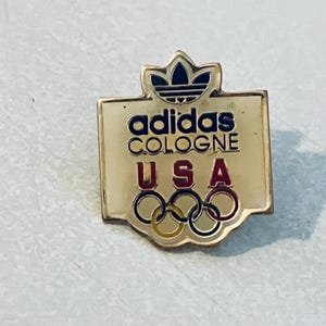 以下が含まれることがあります： 白い背景に、アディダスロゴ、テキスト「adidas COLOGNE」、そして「USA」とオリンピックの輪が描かれた金色のピン。
