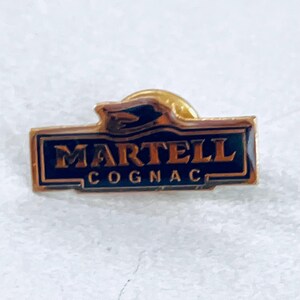May include: A rectangular gold-toned pin with the text "MARTELL COGNAC" in gold lettering on a dark blue background. The pin has a gold border and a small gold emblem at the top.