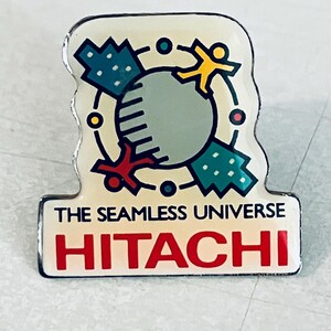 以下が含まれることがあります： 地球を3つの図形が囲むカラフルなグラフィックが描かれた白と黄色のエナメルピン。グラフィックの下に「The Seamless Universe HITACHI」の文字が印刷されています。