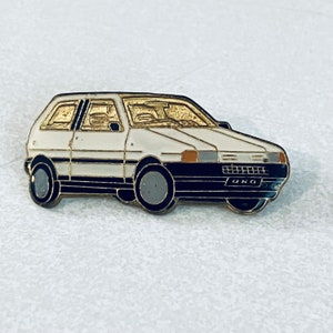 May include: A white Fiat Uno car pin with black trim and gold accents. The car has a black interior and the word "UNO" is visible on the back.