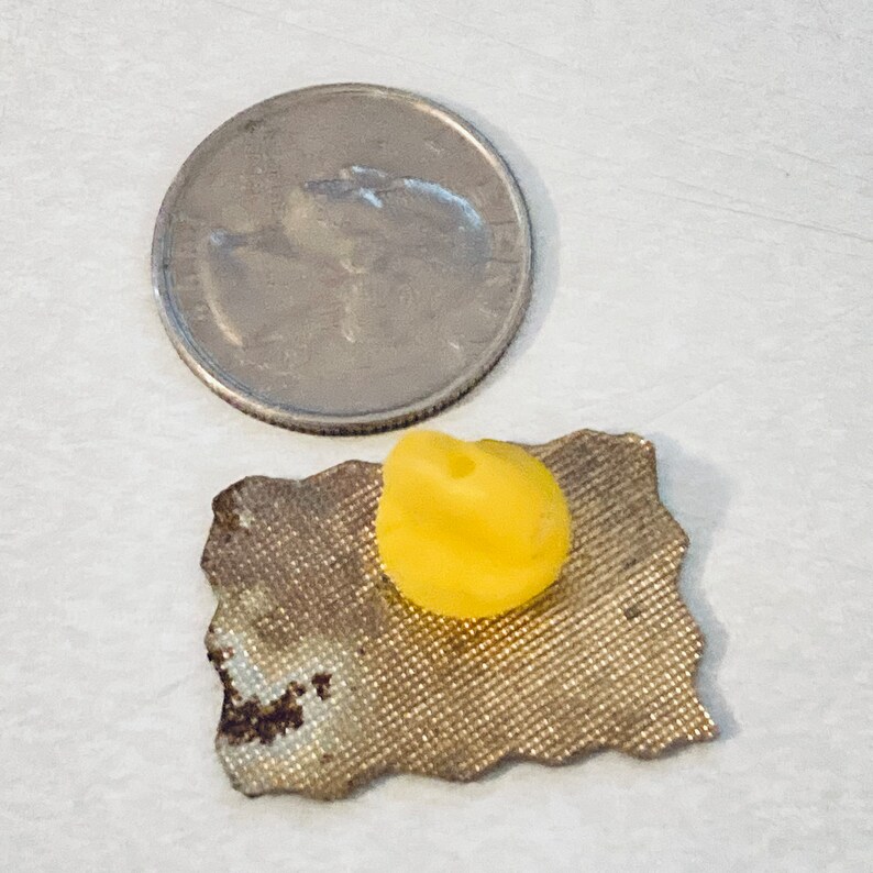 May include: A small, yellow, rubbery object, possibly a button or a cap, is sitting on a piece of brown fabric. The fabric has a textured surface and a slightly irregular shape. A US quarter is shown for scale.