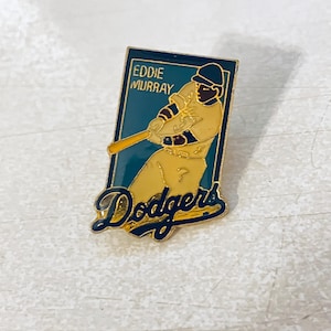 May include: A vintage Dodgers pin featuring a baseball player in a batting stance. The pin has a gold border, a teal background, and the words "EDDIE MURRAY" and "Dodgers" in navy blue. The player is wearing a yellow uniform.
