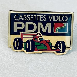 May include: A black and gold pin with the text "CASSETTES VIDEO PDM" and a logo of a blue, green, and red swirl. The pin also features a red race car with a driver.