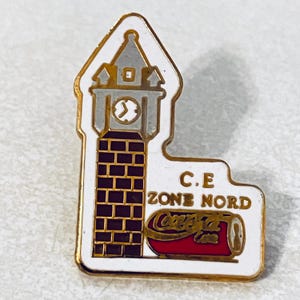 May include: A white and gold enamel pin featuring a clock tower design with the text "C.E ZONE NORD" and a Coca-Cola can. The clock tower has a clock face and a brick pattern. The Coca-Cola can is red and gold.