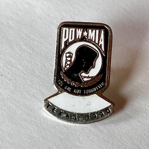 May include: A rectangular POW/MIA pin with a black and white design. The pin features the text "POW*MIA" at the top, a silhouette of a person, and the words "YOU ARE NOT FORGOTTEN". The bottom of the pin reads "TEXAS 1990".