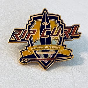 Puede incluir: Un pin metálico dorado con las palabras "RIP CURL" en letras rojas y doradas. El pin presenta un diseño de tabla de surf con el texto "AUSTRALIAN SURFING" en una pancarta. El pin tiene una combinación de colores negro, amarillo y azul.