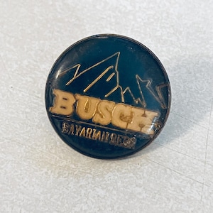 May include: Round, dark blue pin with the word "BUSCH" in tan letters, above a mountain range graphic. The text "BAYARIAN BEER" is below the word "BUSCH".