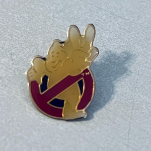 May include: A gold-coloured pin featuring the Ghostbusters logo with a cartoon ghost figure. The ghost is giving a peace sign, and the logo is a red circle with a diagonal line through it. The pin has a shiny finish.
