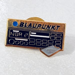 Puede incluir: Un pin de esmalte dorado con forma de radio Blaupunkt vintage. El pin presenta la palabra "BLAUPUNKT" en letras negras, junto con el texto "NDR 2". El pin tiene un círculo azul y un pequeño rectángulo gris.