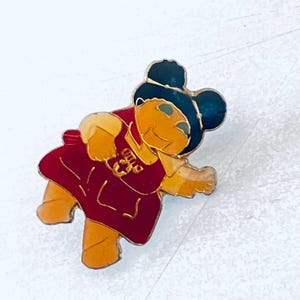 May include: A red and gold enamel pin featuring a cartoon character with black hair, a red dress, and a yellow face. The character is holding a red and gold object in their right hand.