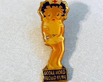 Op de afbeelding: Een vintage speld met een cartoonverpleegster in een gele uniform en muts met een rood kruis. De speld heeft een gouden rand en de tekst "ECOLE NORD PROMO 91/94" op een blauwe rechthoekige basis. De verpleegster heeft donker haar en een rode mond.