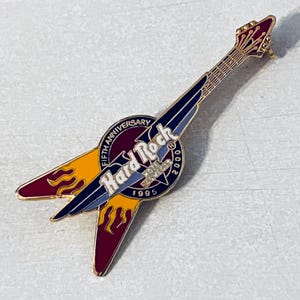 May include: Gold-colored Hard Rock Cafe pin featuring a guitar with flames and the text "Hard Rock Cafe" in a circle with the text "Fifth Anniversary 1995-2000".