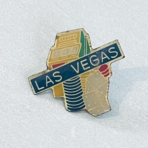 May include: A vintage Las Vegas souvenir pin featuring a colorful design of buildings and landmarks. The pin has a blue banner with the words "LAS VEGAS" in white lettering. The pin is made of metal and has a gold border.