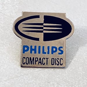 May include: A vintage Philips Compact Disc pin. The pin has a silver-colored metal finish with a blue and black design. The text "PHILIPS COMPACT DISC" is in blue letters.