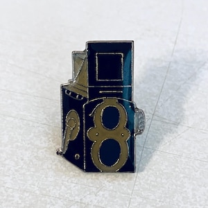 May include: A vintage-style pin featuring a camera design with the number "8" in gold. The pin has a dark blue enamel finish with gold and silver accents. The pin is a collectible item.