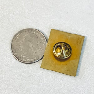 May include: The back of a gold-colored pin with a safety pin closure. The pin is about the size of a US quarter.