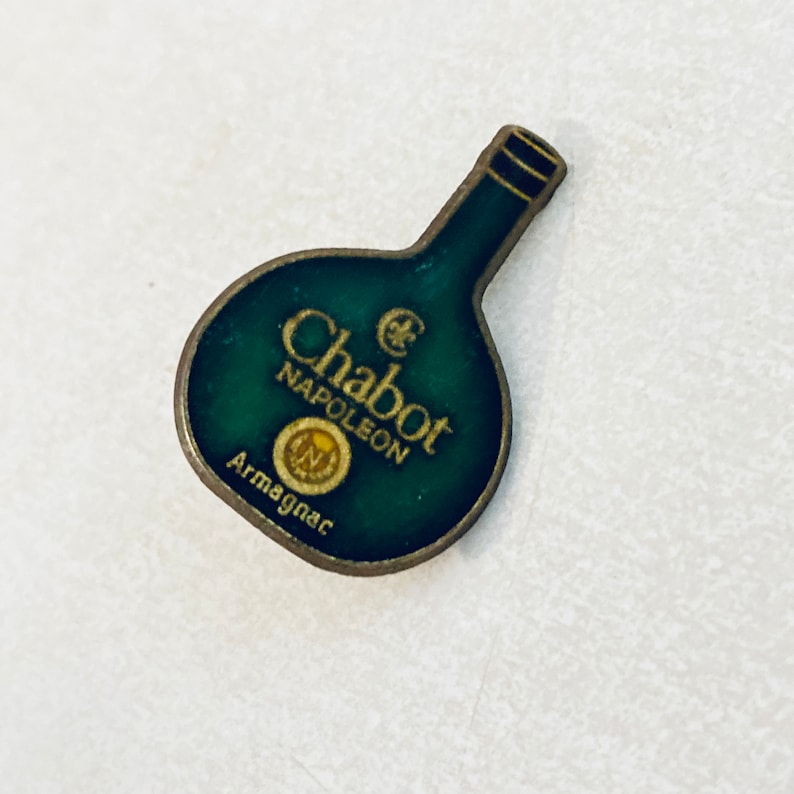 May include: A green and gold metal pin shaped like a bottle of Chabot Napoleon Armagnac.