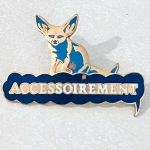 May include: A gold-colored pin with a cartoon fennec fox sitting on a blue cloud with the word "ACCESSOIREMENT" written in white letters.