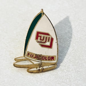 Puede incluir: Un pin de color dorado con forma de velero con una vela blanca, ribete verde y un logotipo "FUJI" rojo y blanco. Las palabras "FUJICOLOR" están impresas en negro en la vela.