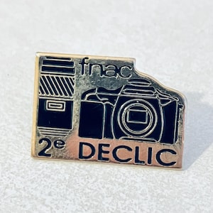 Può includere: Uno spillo in metallo dorato con un'immagine in bianco e nero di una macchina fotografica e un obiettivo. Lo spillo dice "fnac 2e DECLIC".