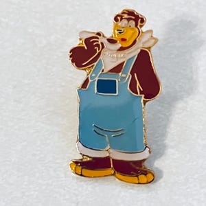 May include: A colorful enamel pin featuring a cartoon bear wearing blue overalls, a white scarf, and brown boots. The bear has a yellow face and is holding a white object to its mouth. The pin has a gold border.