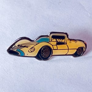 May include: A yellow and black enamel pin shaped like a vintage race car. The pin features a light blue stripe and accents, with dark brown wheels and a black outline. The car has a sleek, aerodynamic design.