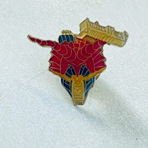 May include: A metal enamel pin featuring a stylized creature head with red, blue, and gold sections. The design includes two prominent horns and a rectangular banner at the top with the text "UNITS" in a bold, embossed, gold-colored font style.